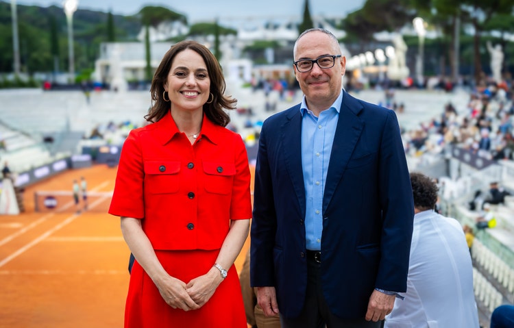 Fotografie von zwei Personen: Marina Storti, CEO von WTA Ventures, und Mauro Macchi, CEO von Accenture EMEA. Beide stehen vor einem Sandplatz‑Tenniscourt, im Hintergrund sind Zuschauer zu sehen. Marina Storti links trägt ein rotes Outfit, Mauro Macchi rechts einen dunklen Anzug mit hellblauem Hemd